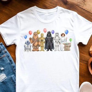 May include: White t-shirt featuring a Star Wars themed graphic. The design includes illustrations of R2-D2, Chewbacca, C-3PO, Darth Vader, a Stormtrooper, Princess Leia, and Grogu, each holding a balloon. The shirt is displayed on a wooden surface.