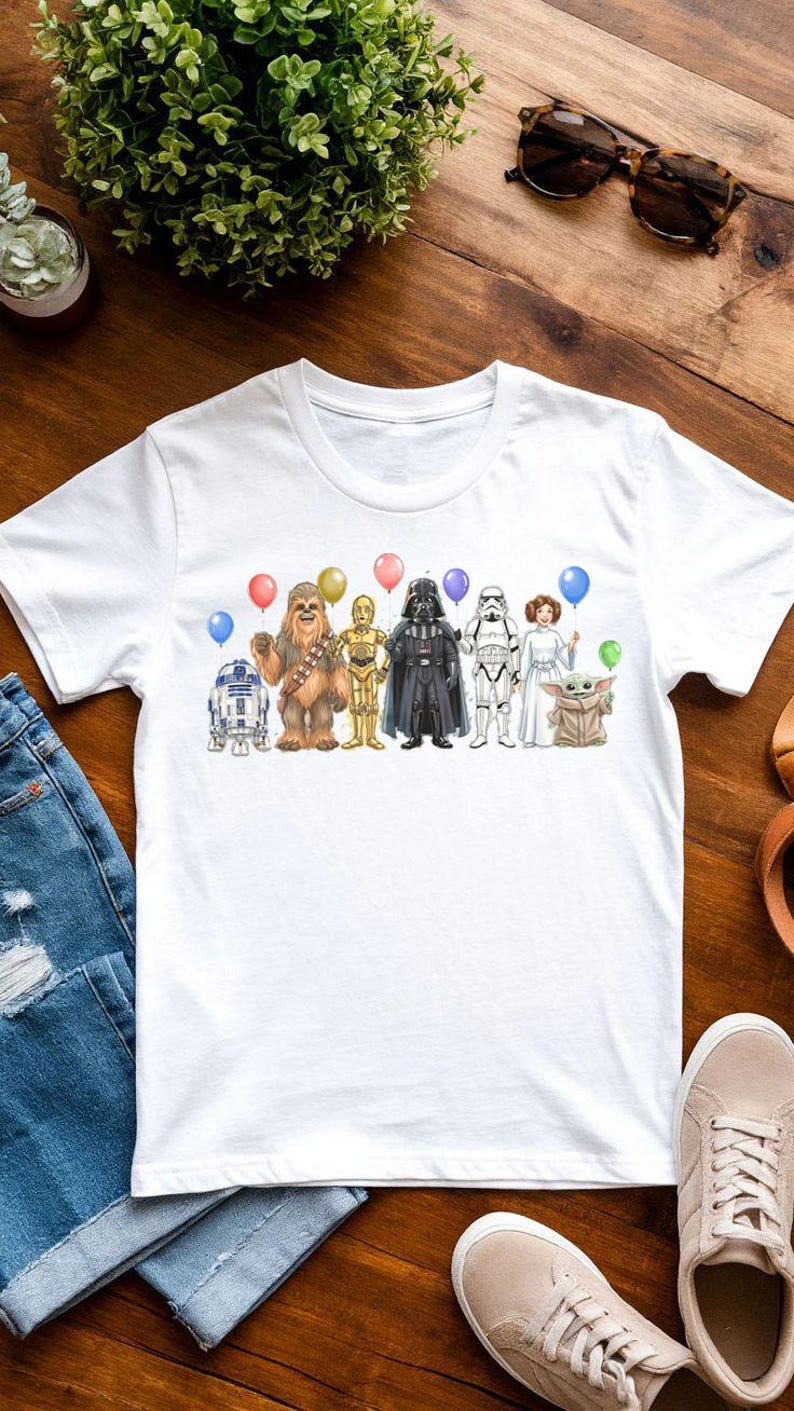 May include: White t-shirt featuring a Star Wars themed graphic. The design includes illustrations of R2-D2, Chewbacca, C-3PO, Darth Vader, a Stormtrooper, Princess Leia, and Grogu, each holding a balloon. The shirt is displayed on a wooden surface.