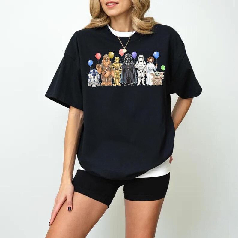 May include: Black t-shirt featuring Star Wars characters holding balloons. The graphic includes R2-D2, Chewbacca, C-3PO, Darth Vader, a Stormtrooper, Princess Leia, and Grogu. The shirt is a casual, oversized fit.