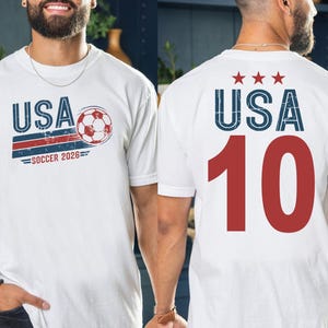 May include: A white t-shirt featuring a graphic on the front with the text USA, a soccer ball, and the text SOCCER 2026. The back of the t-shirt displays the text USA with three red stars above it and the large red number 10 printed below.