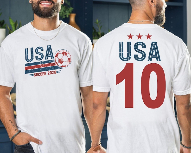 May include: A white t-shirt featuring a graphic on the front with the text USA, a soccer ball, and the text SOCCER 2026. The back of the t-shirt displays the text USA with three red stars above it and the large red number 10 printed below.