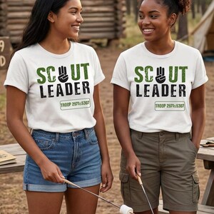 May include: Two women wearing white t-shirts with the text "SCOUT LEADER" in green and black. The shirts also have "TROOP 2971&5307" printed below. One woman wears denim shorts, the other wears olive green cargo shorts. They are holding marshmallow roasting sticks.