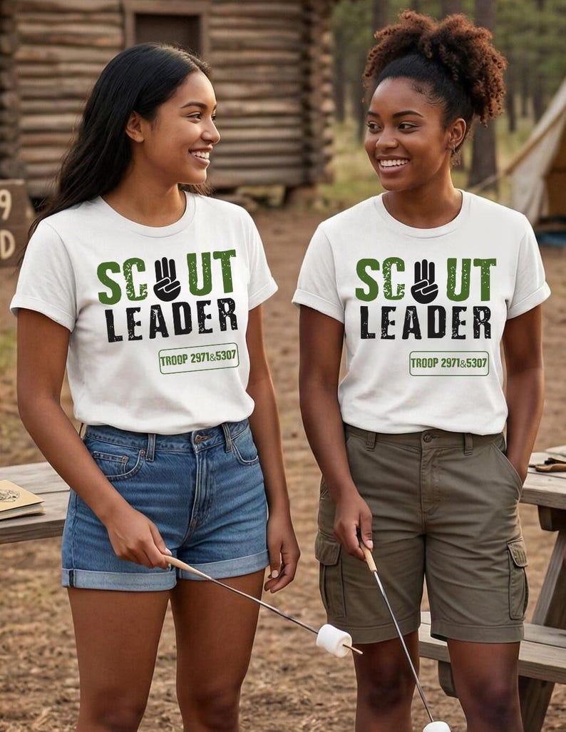 May include: Two women wearing white t-shirts with the text "SCOUT LEADER" in green and black. The shirts also have "TROOP 2971&5307" printed below. One woman wears denim shorts, the other wears olive green cargo shorts. They are holding marshmallow roasting sticks.
