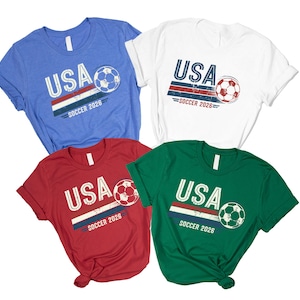 May include: Four short-sleeve t-shirts in blue, white, red, and green. Each shirt features a graphic with the text USA and SOCCER 2026, accompanied by a soccer ball illustration and horizontal stripes in a distressed, vintage-inspired print style.