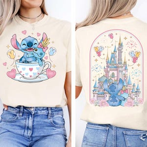 Puede incluir: Camiseta color crema con una ilustraci&oacute;n de dibujos animados de Stitch y Angel en una taza de t&eacute;, rodeados de corazones y globos. La parte posterior de la camiseta muestra a Stitch y Angel frente a un castillo con fuegos artificiales.