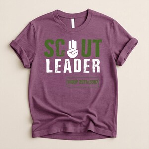 May include: A purple t-shirt with rolled-up sleeves. The shirt features the words "SCOUT LEADER" in green and white block letters, with a hand symbol. Below the text is the text "TROOP 2971 & 5307" in a green rectangle.