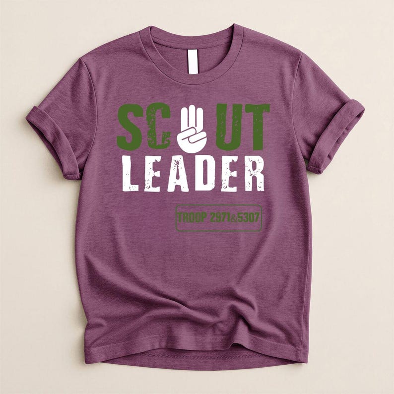 May include: A purple t-shirt with rolled-up sleeves. The shirt features the words "SCOUT LEADER" in green and white block letters, with a hand symbol. Below the text is the text "TROOP 2971 & 5307" in a green rectangle.