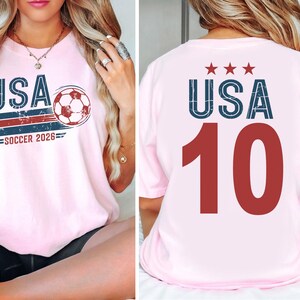 May include: A light pink t-shirt featuring a front graphic with the text USA, a soccer ball, and the text SOCCER 2026. The back of the t-shirt displays the text USA with three stars above it and the large red number 10 printed in the center.