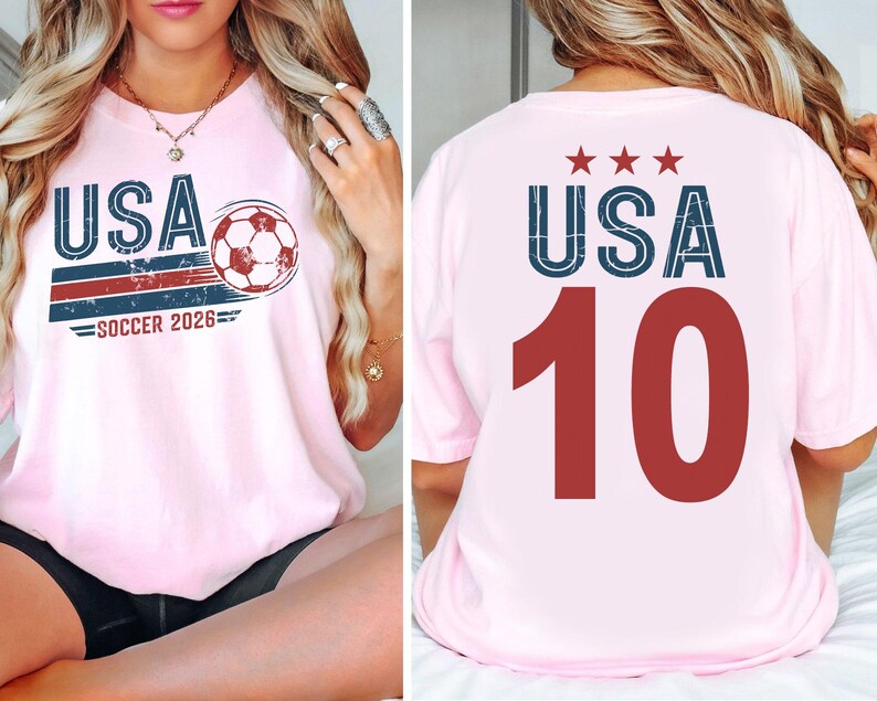 May include: A light pink t-shirt featuring a front graphic with the text USA, a soccer ball, and the text SOCCER 2026. The back of the t-shirt displays the text USA with three stars above it and the large red number 10 printed in the center.