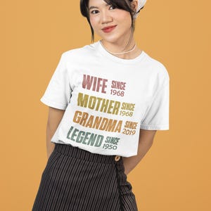 May include: A white t-shirt with the words "WIFE SINCE 1968, MOTHER SINCE 1968, GRANDMA SINCE 2019, LEGEND SINCE 1950" in a colorful, vintage font. The person is wearing a black and white striped skirt and a white bandana.