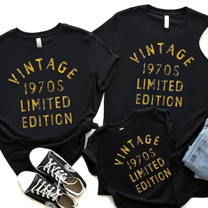 May include: Black t-shirts with the text "VINTAGE 1970s LIMITED EDITION" in yellow. The image includes a pair of black sneakers, a white baseball cap, and a pair of blue jeans. The shirts are likely for a family or group.
