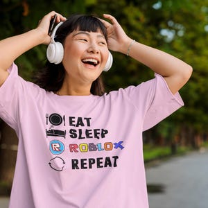 Puede incluir: Una camiseta rosa claro con el texto "EAT SLEEP ROBLOX REPEAT" en negro, azul, rojo y amarillo. La camiseta presenta iconos gr&aacute;ficos de un plato y un tenedor, una cama, el logo de Roblox y un s&iacute;mbolo de reciclaje. La persona lleva auriculares blancos.