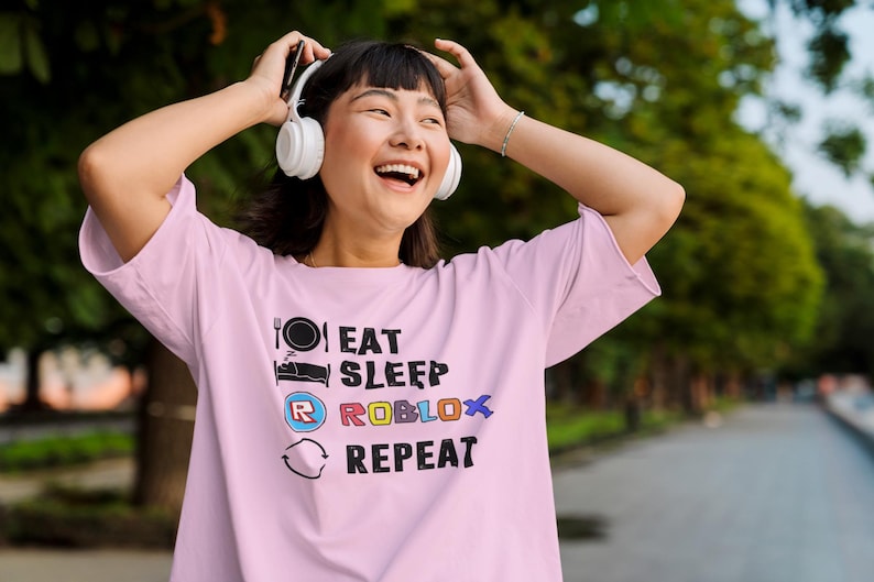 Puede incluir: Una camiseta rosa claro con el texto "EAT SLEEP ROBLOX REPEAT" en negro, azul, rojo y amarillo. La camiseta presenta iconos gr&aacute;ficos de un plato y un tenedor, una cama, el logo de Roblox y un s&iacute;mbolo de reciclaje. La persona lleva auriculares blancos.