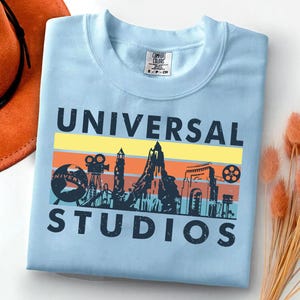 May include: Light blue crewneck sweatshirt with the words "UNIVERSAL STUDIOS" in black block letters. The design features a colorful retro graphic of the Universal Studios logo with a film reel, camera, and mountain silhouette.