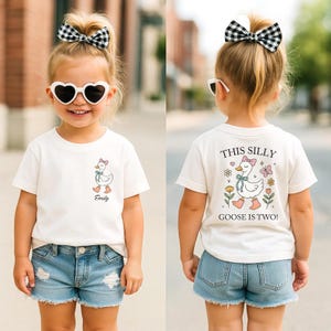 May include: A cream-colored t-shirt with a cartoon goose design and the name "Emily" printed below it. The back of the shirt says "This Silly Goose is Two!" with a goose illustration and floral accents. The child is wearing denim shorts and heart-shaped sunglasses.
