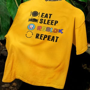 Puede incluir: Camiseta de color amarillo mostaza con las palabras "EAT SLEEP ROBLOX REPEAT" impresas en negro, azul y naranja. La camiseta presenta iconos de un plato y un tenedor, una cama, el logotipo de Roblox y una flecha circular.