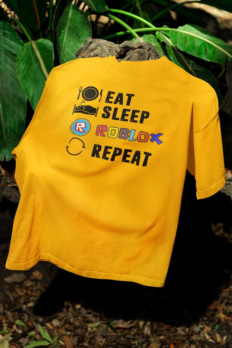 Puede incluir: Camiseta de color amarillo mostaza con las palabras "EAT SLEEP ROBLOX REPEAT" impresas en negro, azul y naranja. La camiseta presenta iconos de un plato y un tenedor, una cama, el logotipo de Roblox y una flecha circular.