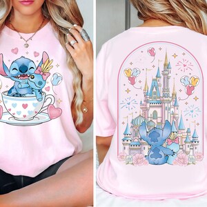 Puede incluir: Camiseta rosa claro con un personaje de dibujos animados Stitch y una mu&ntilde;eca Stitch m&aacute;s peque&ntilde;a en una taza de t&eacute; decorada con corazones. La parte trasera de la camiseta muestra a Stitch y una mu&ntilde;eca frente a un castillo con fuegos artificiales y globos.