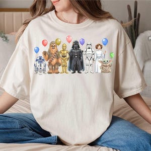 May include: Cream-colored t-shirt featuring a colorful illustration of Star Wars characters holding balloons. The design includes R2-D2, Chewbacca, C-3PO, Darth Vader, a Stormtrooper, Princess Leia, and Grogu.