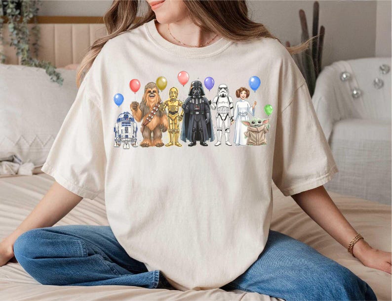 May include: Cream-colored t-shirt featuring a colorful illustration of Star Wars characters holding balloons. The design includes R2-D2, Chewbacca, C-3PO, Darth Vader, a Stormtrooper, Princess Leia, and Grogu.