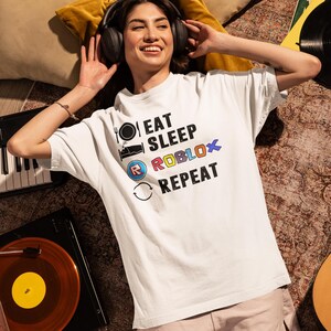 Puede incluir: Camiseta blanca con el texto "EAT SLEEP ROBLOX REPEAT" en negro, azul y rojo. La camiseta presenta iconos de tenedor y plato, una cama y una flecha circular. La persona lleva auriculares y est&aacute; tumbada en una alfombra estampada.