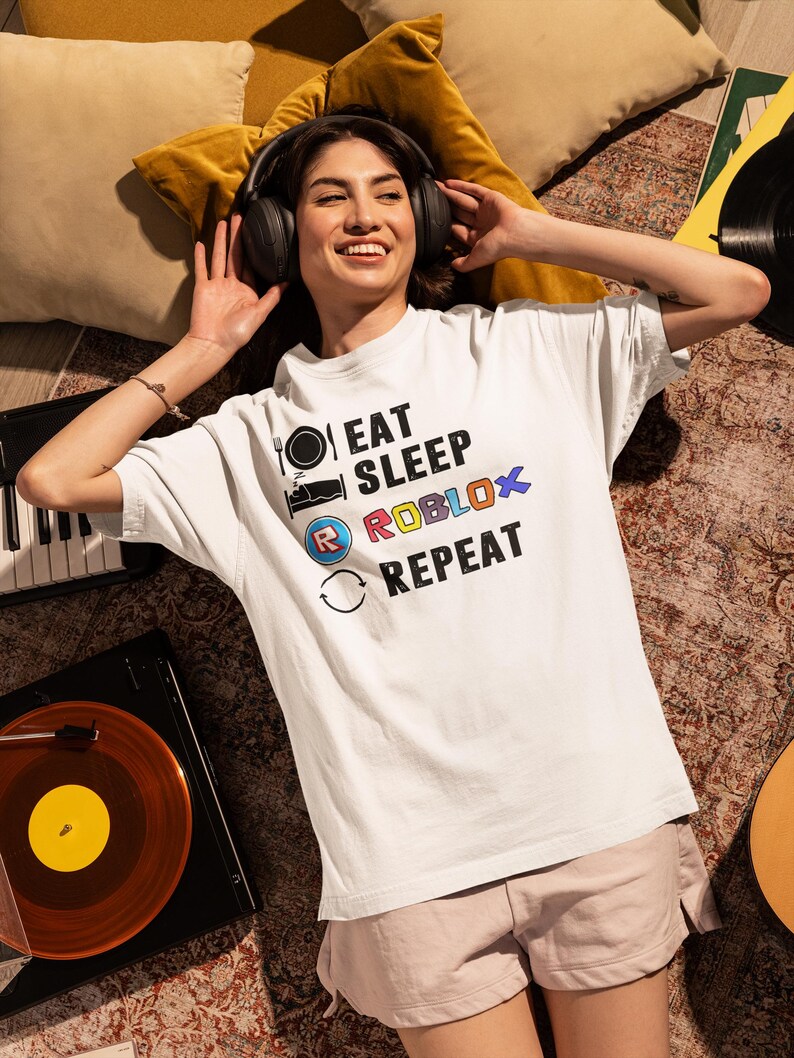 Puede incluir: Camiseta blanca con el texto "EAT SLEEP ROBLOX REPEAT" en negro, azul y rojo. La camiseta presenta iconos de tenedor y plato, una cama y una flecha circular. La persona lleva auriculares y est&aacute; tumbada en una alfombra estampada.