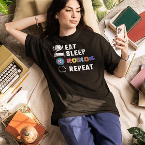 Puede incluir: Una camiseta negra con el texto "EAT SLEEP ROBLOX REPEAT" en letras blancas y coloridas. La persona lleva pantalones azules y est&aacute; acostada en una cama con almohadas, libros y una m&aacute;quina de escribir. La persona sostiene un tel&eacute;fono.