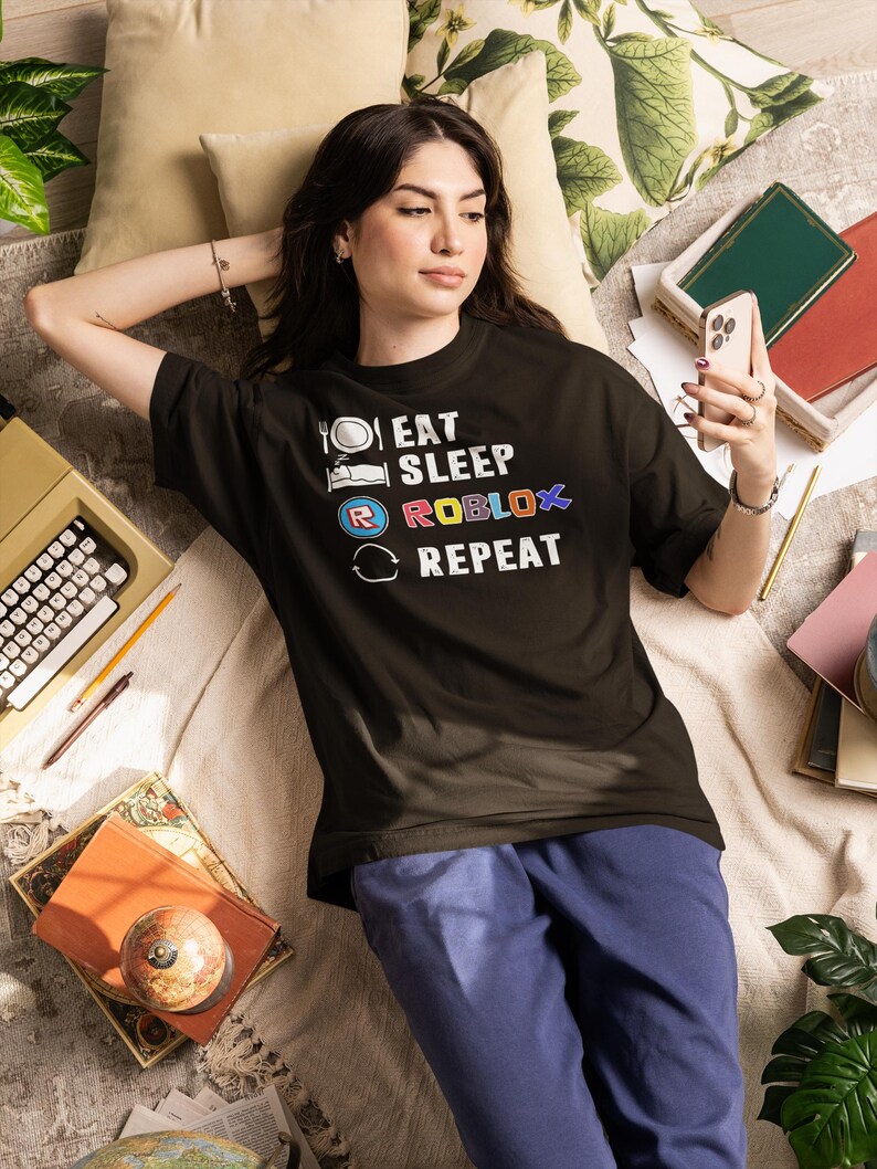 Puede incluir: Una camiseta negra con el texto "EAT SLEEP ROBLOX REPEAT" en letras blancas y coloridas. La persona lleva pantalones azules y est&aacute; acostada en una cama con almohadas, libros y una m&aacute;quina de escribir. La persona sostiene un tel&eacute;fono.