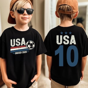 May include: A black t-shirt featuring a front graphic with the text USA, a soccer ball, and the text SOCCER 2026 above red, white, and blue stripes. The back of the shirt displays the text USA with three stars above and the large blue number 10.