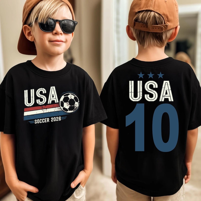 May include: A black t-shirt featuring a front graphic with the text USA, a soccer ball, and the text SOCCER 2026 above red, white, and blue stripes. The back of the shirt displays the text USA with three stars above and the large blue number 10.