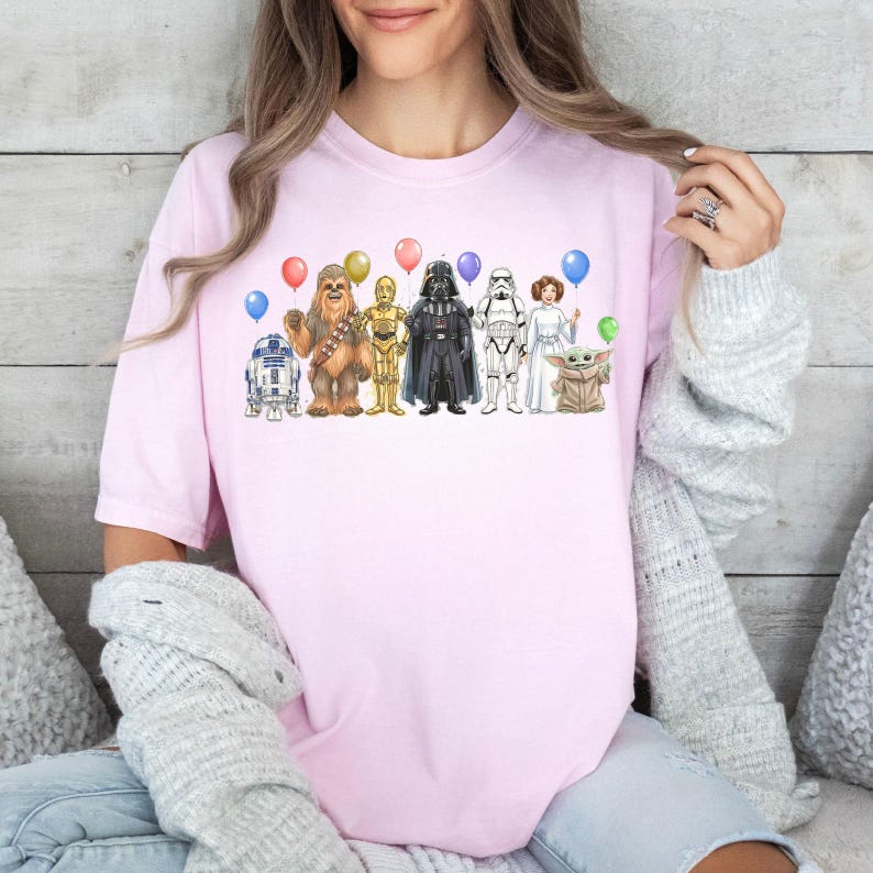 May include: A light pink t-shirt featuring a Star Wars-themed graphic. The graphic includes illustrations of various characters such as R2-D2, Chewbacca, C-3PO, Darth Vader, a Stormtrooper, Princess Leia, and Baby Yoda, each holding a balloon.