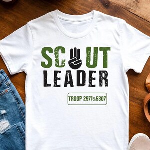 May include: White t-shirt with the words "SCOUT LEADER" in green and black block letters. A black hand symbol is between the words "SCOUT" and "LEADER". The text "TROOP 2971&5307" is below the words. The shirt is on a wooden surface.