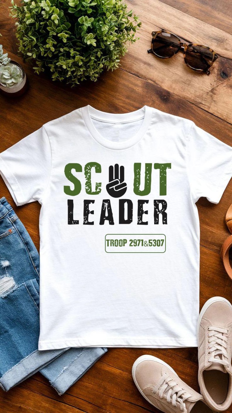May include: White t-shirt with the words "SCOUT LEADER" in green and black block letters. A black hand symbol is between the words "SCOUT" and "LEADER". The text "TROOP 2971&5307" is below the words. The shirt is on a wooden surface.