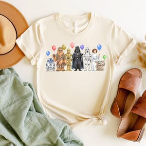 May include: Cream-colored t-shirt featuring a colorful illustration of Star Wars characters, including R2-D2, Chewbacca, C-3PO, Darth Vader, a Stormtrooper, Princess Leia, and Grogu, each holding a balloon. The shirt is paired with brown sandals and a tan hat.