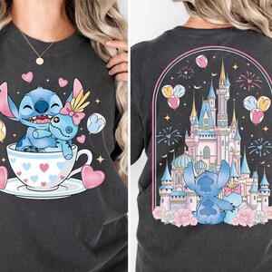 Puede incluir: Camiseta gris oscuro con un dibujo animado de Stitch y una mu&ntilde;eca en una taza de t&eacute; con corazones. La parte trasera de la camiseta muestra a Stitch y la mu&ntilde;eca frente a un castillo con fuegos artificiales y globos. El dise&ntilde;o es en tonos azules, rosas y amarillos.