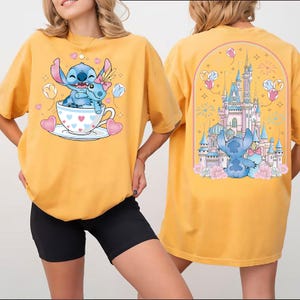 Puede incluir: Camiseta amarilla mostaza con un dise&ntilde;o de dibujos animados de Stitch. La parte delantera muestra a Stitch en una taza de t&eacute; con corazones y globos. La parte trasera muestra a Stitch con un castillo y fuegos artificiales. Una camiseta divertida e informal.