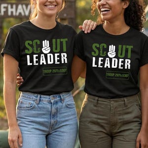 May include: Two people wearing black t-shirts with the words "SCOUT LEADER" in white and green lettering. The shirts also have "TROOP 2971&5307" printed below the text. One person is wearing blue jeans, and the other is wearing olive green pants.