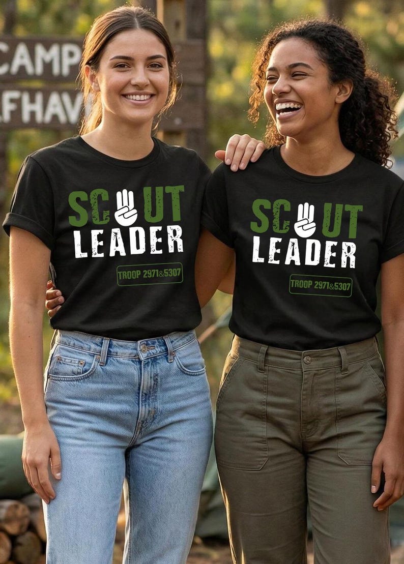 May include: Two people wearing black t-shirts with the words "SCOUT LEADER" in white and green lettering. The shirts also have "TROOP 2971&5307" printed below the text. One person is wearing blue jeans, and the other is wearing olive green pants.