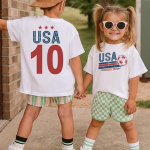May include: Two white t-shirts featuring graphic prints. One shirt displays the text USA with three stars and the number 10 in red. The other shirt features the text USA SOCCER 2026 with a soccer ball graphic. Both shirts are worn with patterned shorts.