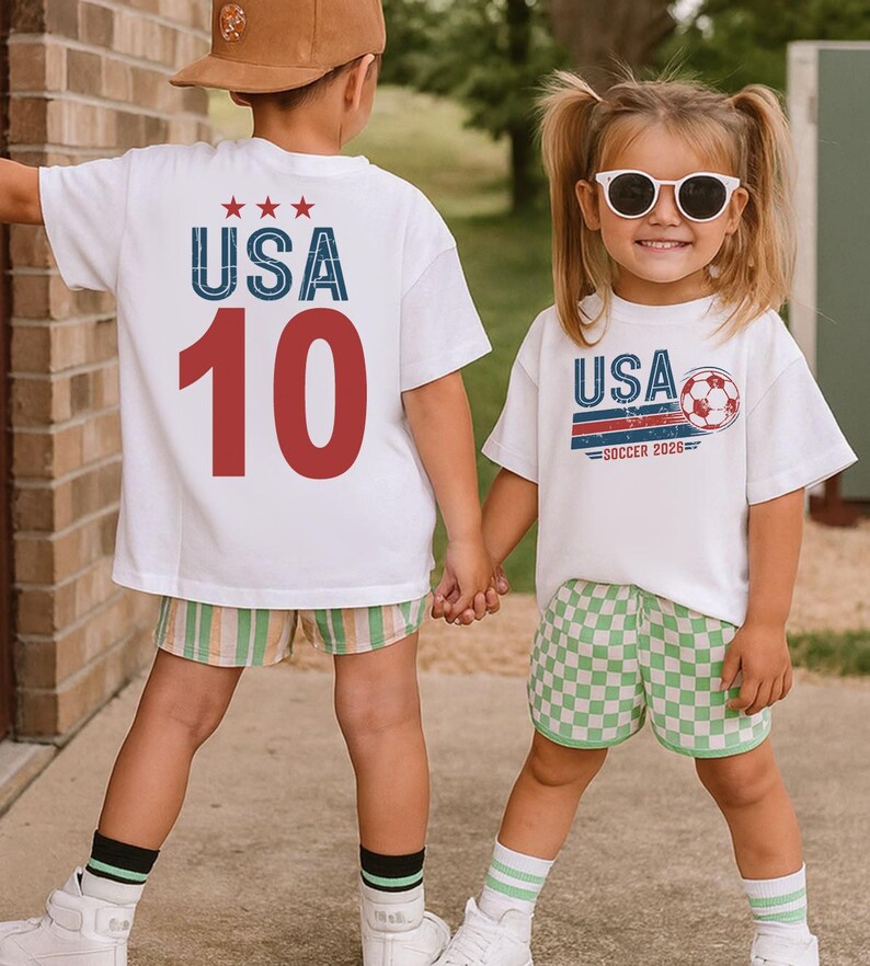 May include: Two white t-shirts featuring graphic prints. One shirt displays the text USA with three stars and the number 10 in red. The other shirt features the text USA SOCCER 2026 with a soccer ball graphic. Both shirts are worn with patterned shorts.