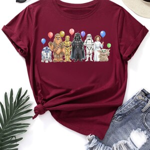May include: Burgundy t-shirt featuring a colorful graphic of Star Wars characters holding balloons. The design includes R2-D2, Chewbacca, C-3PO, Darth Vader, a Stormtrooper, Princess Leia, and Grogu. The shirt is paired with denim shorts and sunglasses.