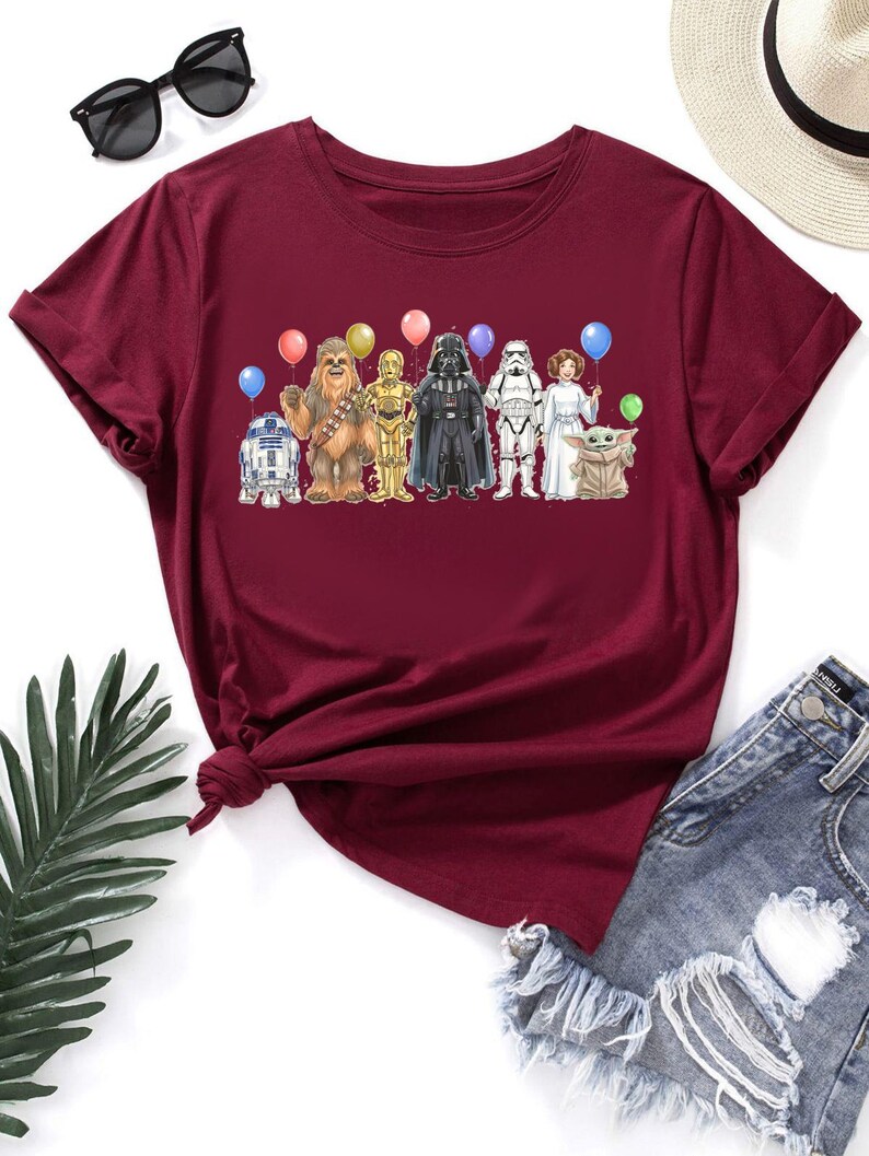 May include: Burgundy t-shirt featuring a colorful graphic of Star Wars characters holding balloons. The design includes R2-D2, Chewbacca, C-3PO, Darth Vader, a Stormtrooper, Princess Leia, and Grogu. The shirt is paired with denim shorts and sunglasses.
