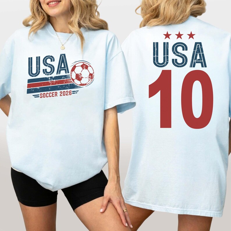 May include: A light blue t-shirt featuring a front graphic with the text USA, a soccer ball, and SOCCER 2026 in red, white, and blue. The back of the shirt displays the text USA with three red stars above the large red number 10 in a distressed font.