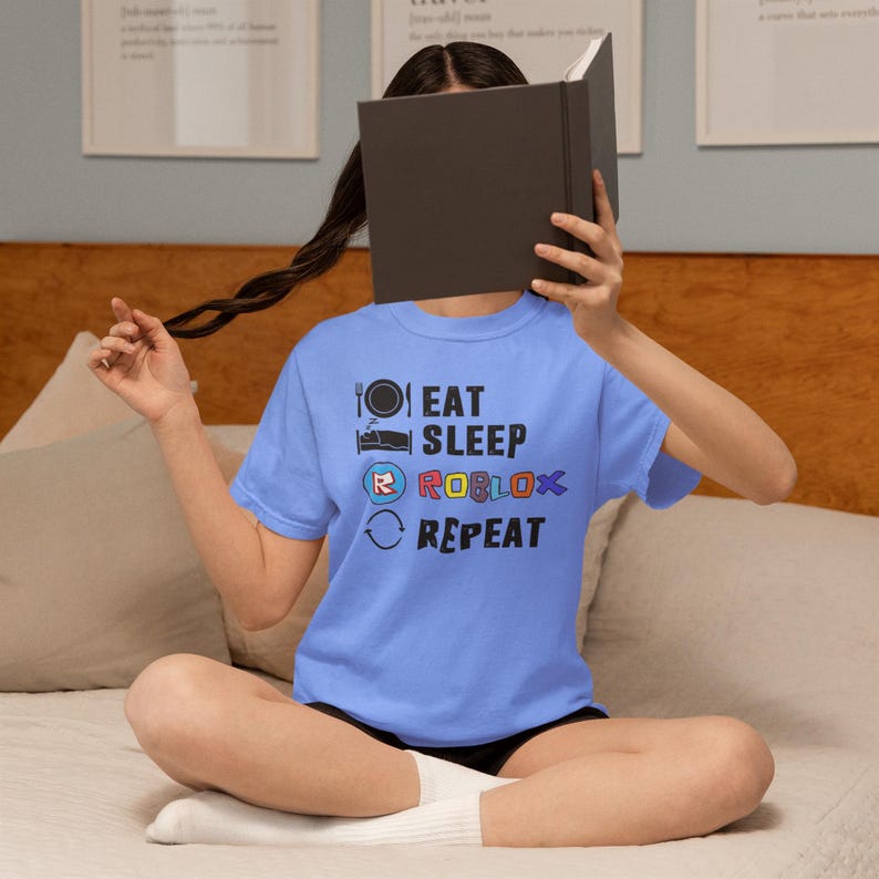 Puede incluir: Una camiseta azul claro con la frase "EAT SLEEP ROBLOX REPEAT" impresa en negro, rojo, azul y amarillo. La camiseta de manga corta la lleva una persona.