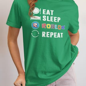 Puede incluir: Camiseta verde con el texto blanco "EAT SLEEP ROBLOX REPEAT", con acentos coloridos en la palabra "ROBLOX". El dise&ntilde;o incluye iconos que representan comer, dormir y el logotipo de Roblox. La camiseta tiene cuello redondo y mangas cortas.