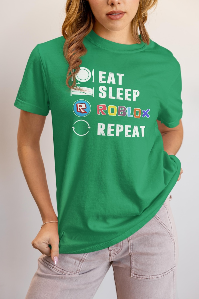 Puede incluir: Camiseta verde con el texto blanco "EAT SLEEP ROBLOX REPEAT", con acentos coloridos en la palabra "ROBLOX". El dise&ntilde;o incluye iconos que representan comer, dormir y el logotipo de Roblox. La camiseta tiene cuello redondo y mangas cortas.