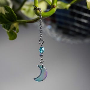 Peut inclure: Une chaîne en argent avec un pendentif en forme de croissant de lune et des perles bleues et argentées suspendues à une plante.