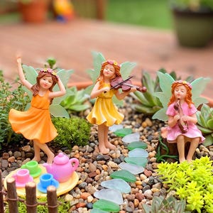 May include: A terracotta planter containing a miniature garden with three fairy figurines, a small wooden fence, a path of green glass stones, succulents, and a tea set. One fairy wears orange, one yellow, and one pink, with small mushrooms nearby.