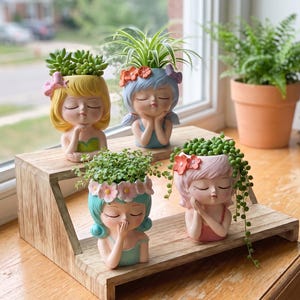 Peut inclure: Quatre pots de fleurs en résine en forme de tête disposés sur un support en bois à deux niveaux. Chaque pot présente un visage aux yeux fermés avec des cheveux de couleurs distinctes : jaune, bleu, turquoise et rose, contenant des plantes.