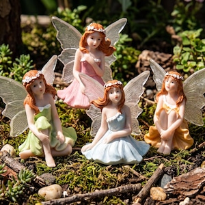 May include: Four small fairy figurines with auburn hair and translucent wings, each wearing a floral crown and a dress in a different pastel color: light green, pink, light blue, and yellow, all posed in a seated position on the ground.